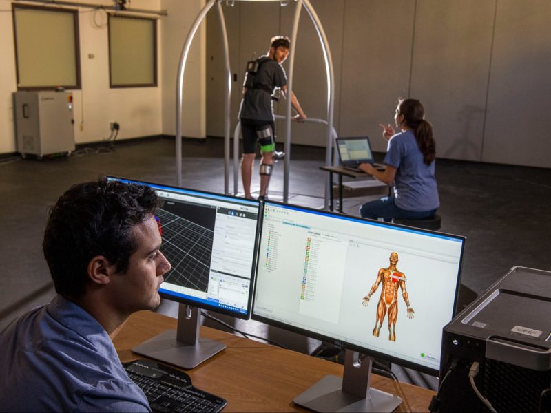 Two people working on computers, a person standing on a raised platform, and a computer screen displaying a 3D human anatomy model.