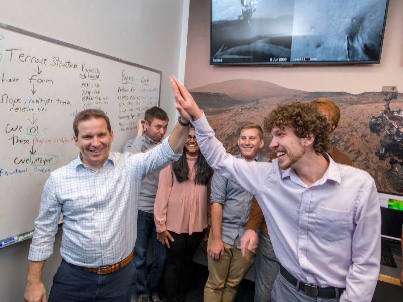 Several people standing in an office setting, smiling and interacting with each other. A whiteboard with writing on it is visible in the background.