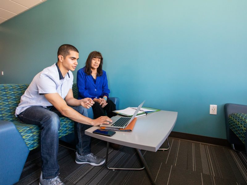 Two people sitting at a table, working on a laptop computer. The individuals appear to be engaged in a conversation or collaborative task. The setting is a modern, colorful office space with teal walls and patterned flooring.