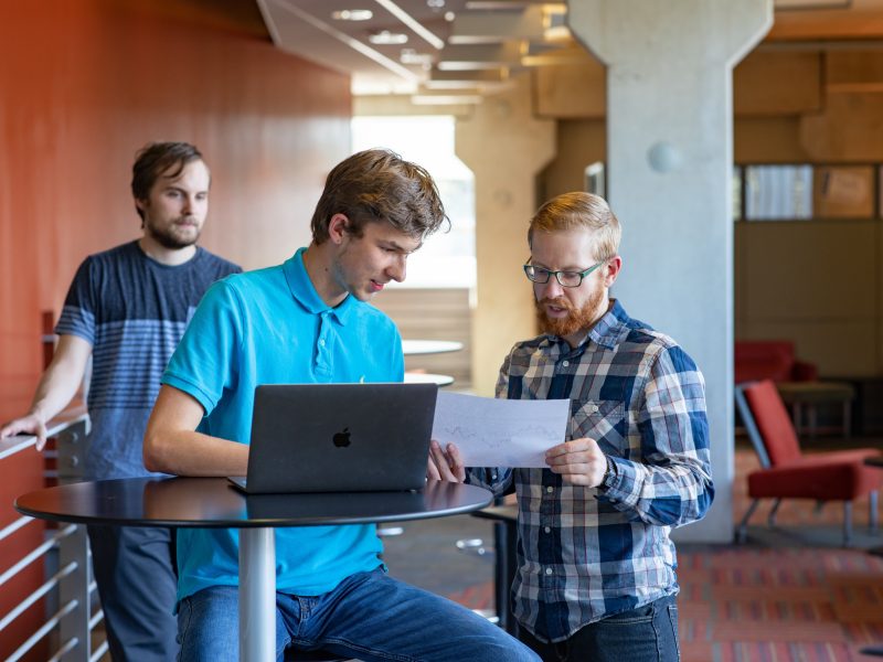 Three people in a casual office setting, one using a laptop and another reviewing documents, with a third person standing in the background.