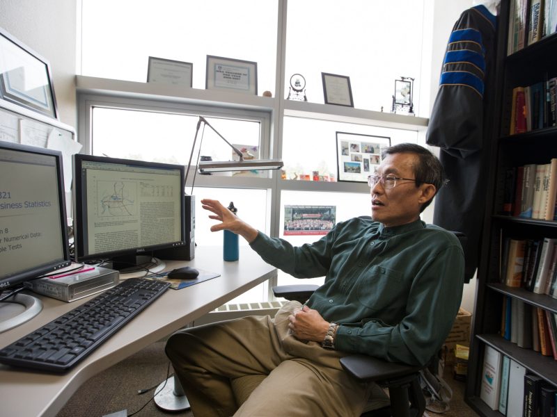 A person sitting at a desk in an office or study area. The person is using a computer and appears to be doing research or academic work, surrounded by bookshelves and other academic materials. The person is wearing casual clothing and appears to be middle-aged or older.