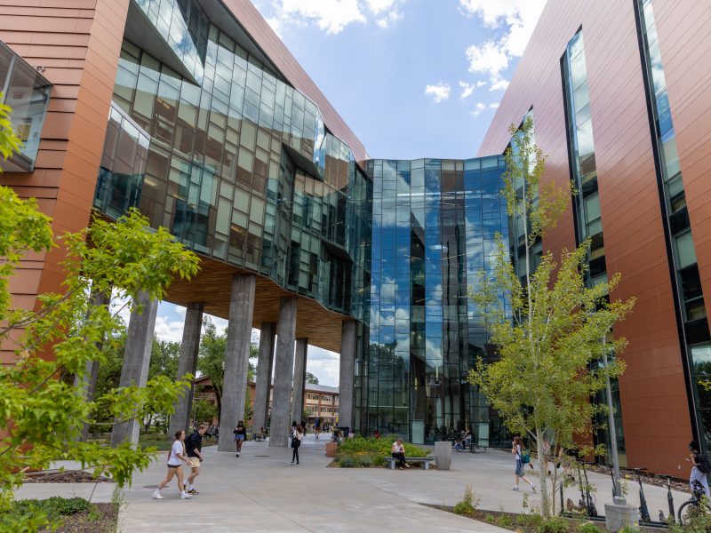 Exterior of a modern multi-story building with glass walls and wooden accents, surrounded by trees and people walking along the sidewalk.