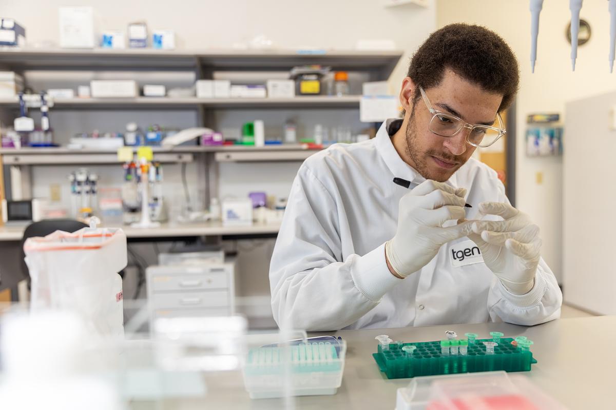Person wearing a white lab coat and gloves, working in a laboratory setting. The person appears to be handling scientific equipment and materials. The laboratory contains various shelves and equipment.