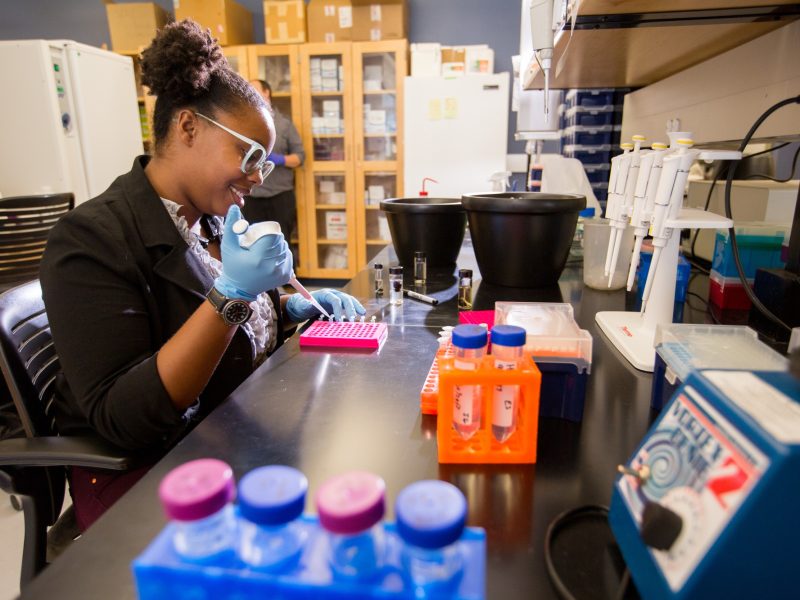 Person wearing lab coat, goggles, and gloves working with scientific equipment and materials in a laboratory setting. Shelves, containers, and other lab equipment visible in the background.