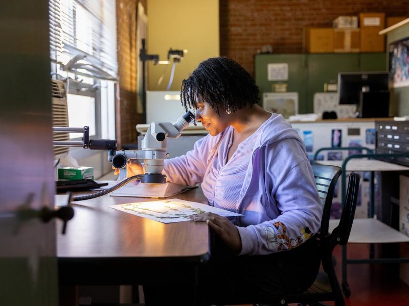 Woman working at desk in office, using laptop computer and writing on papers. Books, files, and other office equipment visible in the background.