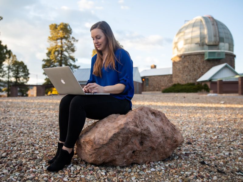 Person using a laptop computer while sitting on a large rock outdoors, with a domed observatory building visible in the background.
