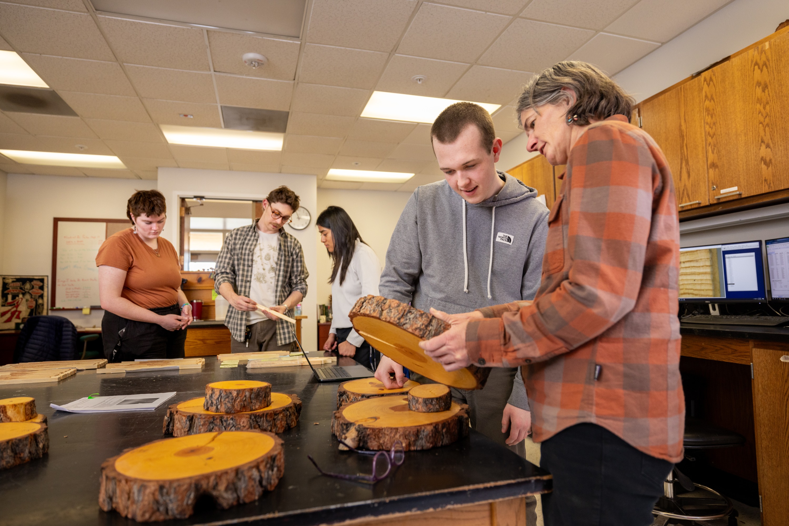 Classroom setting with several people examining wooden logs placed on a table. Some people are standing around the table, while others are seated. The people appear to be engaged in an activity involving the wooden logs.