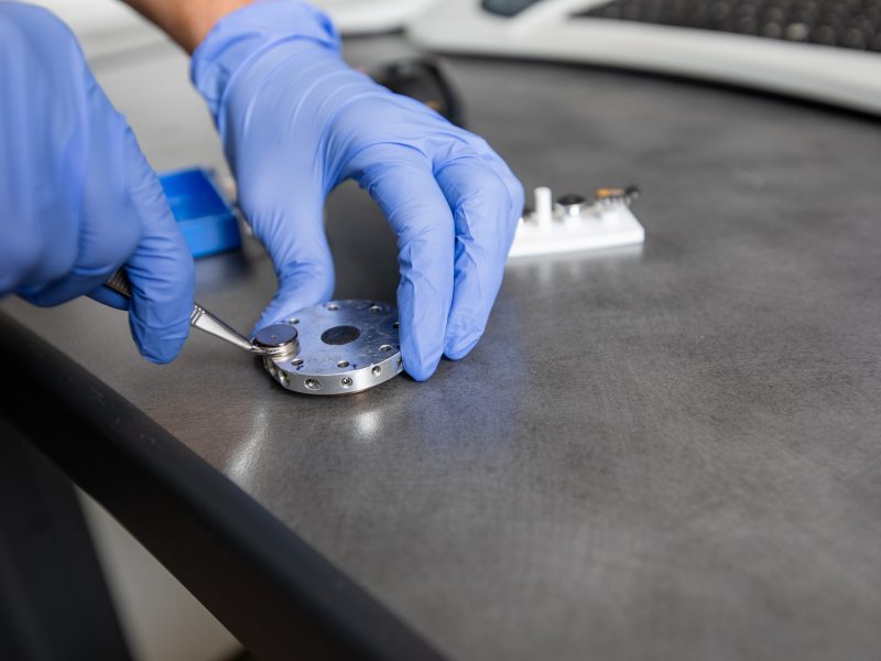 Hands wearing blue gloves holding a small metal component on a dark gray surface, with a keyboard visible in the background.