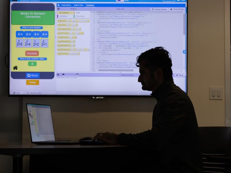 Silhouetted figure of a person sitting at a computer, with a large screen displaying a software program interface.