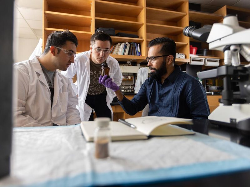 Three individuals in white lab coats and casual attire standing around a table and reviewing documents. They appear to be engaged in discussion or analysis, with a microscope and other scientific equipment visible in the background.