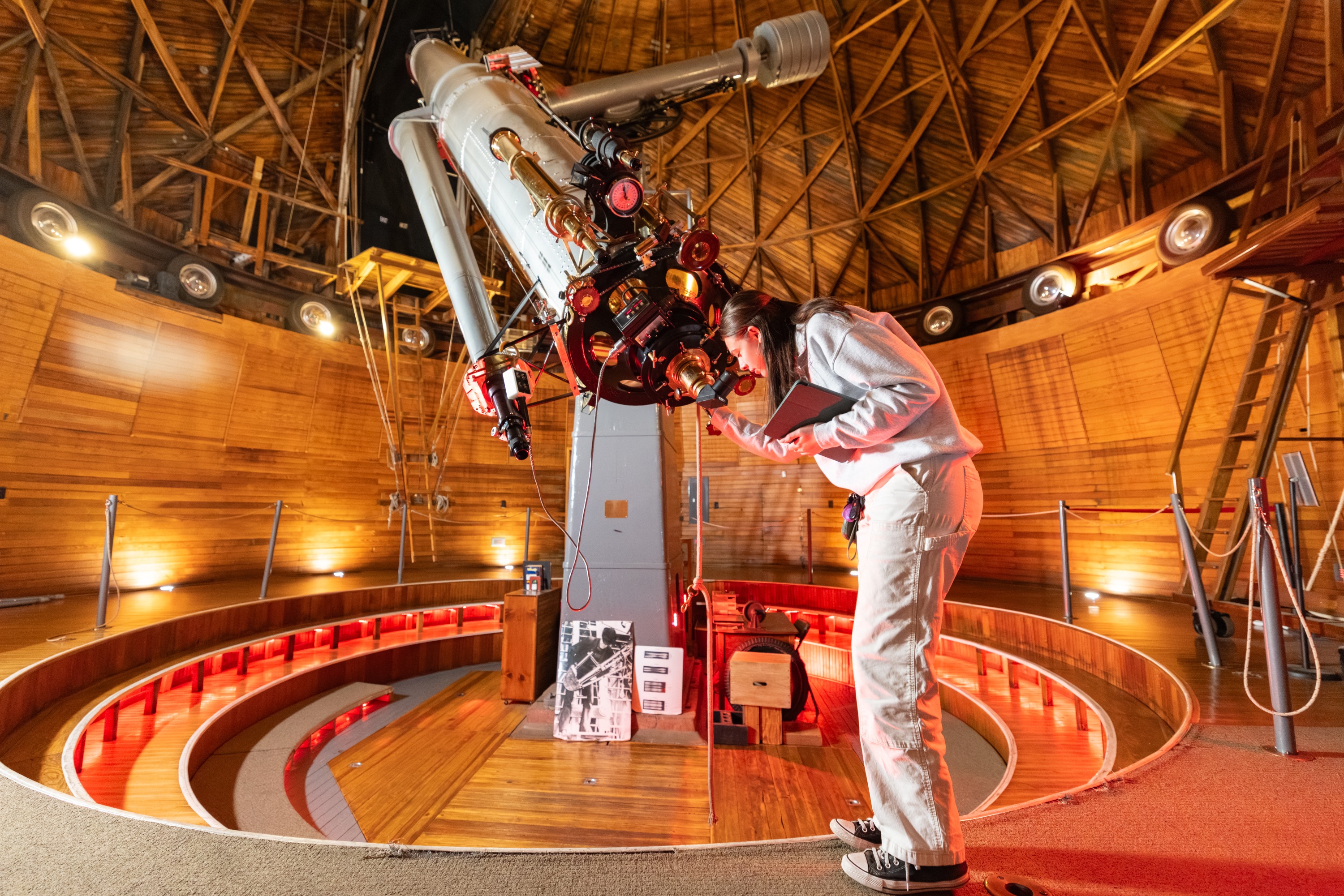 A person wearing a white protective suit, observing a large telescope located in a dome-like structure with wooden beams and red circular seating. The telescope appears to be the main focus of the image, with various equipment and instruments surrounding it.