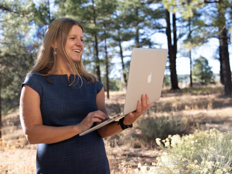 A person standing outdoors, holding a laptop computer and smiling. The person is wearing a navy blue shirt and appears to be in a forest setting. There are pine trees visible in the background.