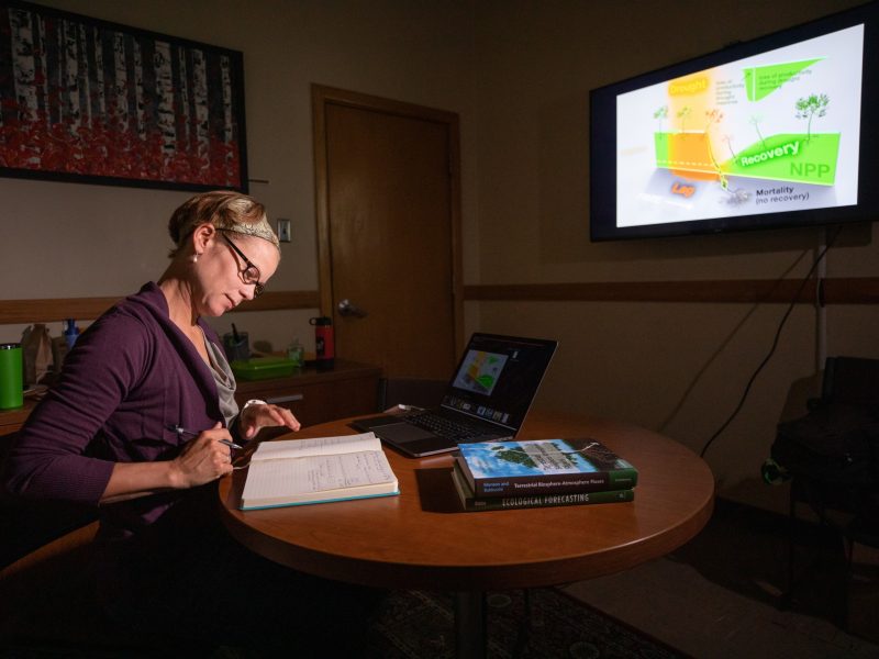 Seated person at a wooden table, working on a laptop computer. Television monitor displays a map and graphics. Books, papers, and office supplies surround the work area.