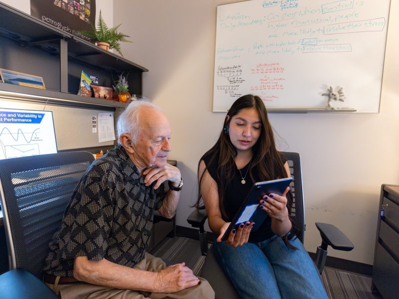 Elderly person with white hair sitting in a chair, and a younger person with dark hair sitting on a chair next to them. There is a whiteboard visible in the background.
