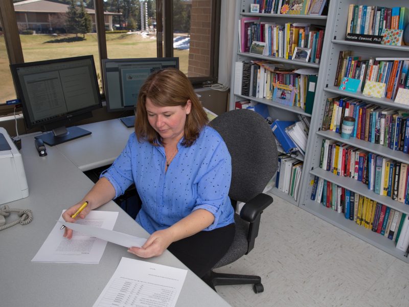 A person sitting at a desk in an office. The person is wearing a blue and white patterned shirt and appears to be reviewing or working on some documents or papers. The background includes bookshelves filled with books and other office equipment.
