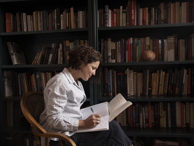 A person in a white shirt sitting in a chair, reading a book in a room filled with bookshelves.
