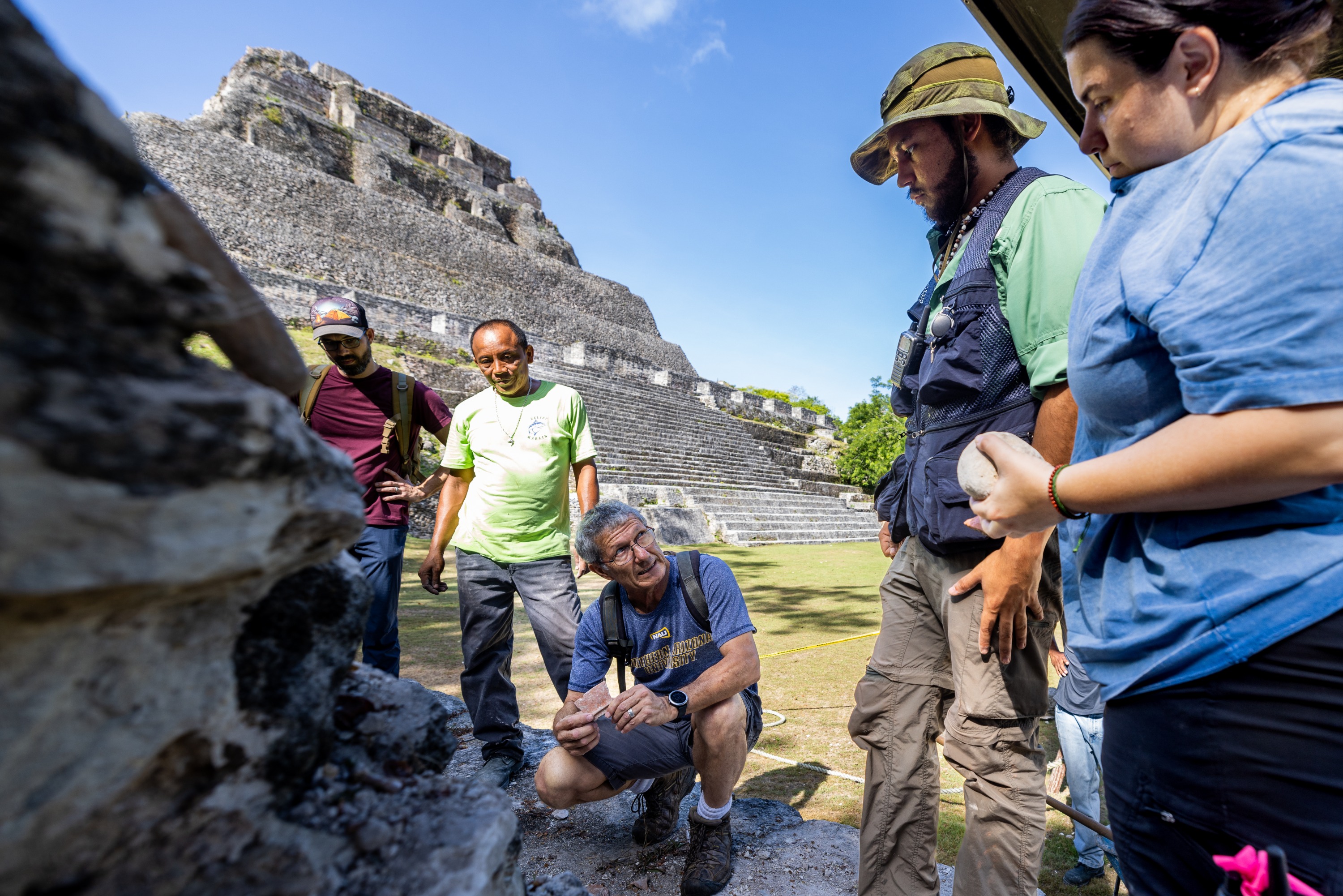 A group of people standing in front of a large stone structure, appearing to be an archaeological site or ancient ruin. The people are wearing casual clothing and exploring the area.