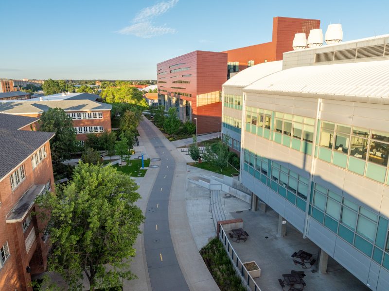 Aerial view of the N A U campus, featuring modern multi-story buildings, a paved walkway, trees, and a clear blue sky.