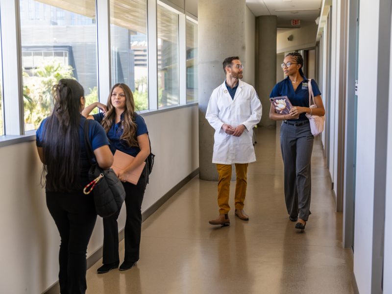 Four people standing in a hallway. Two are wearing white coats, one is wearing a blue uniform, and one is wearing a black uniform. They appear to be in a medical or healthcare setting.