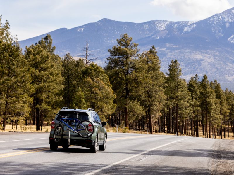 SUV with mountain bikes attached, on a road surrounded by pine trees and mountains in the background.