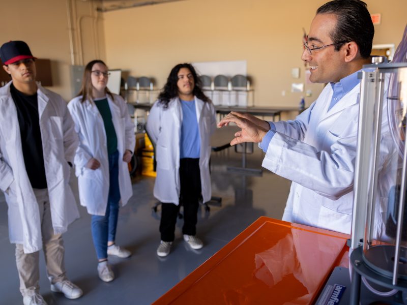 Four individuals in white lab coats, one of whom is pointing at an object on a table. They appear to be in a laboratory or research facility setting.