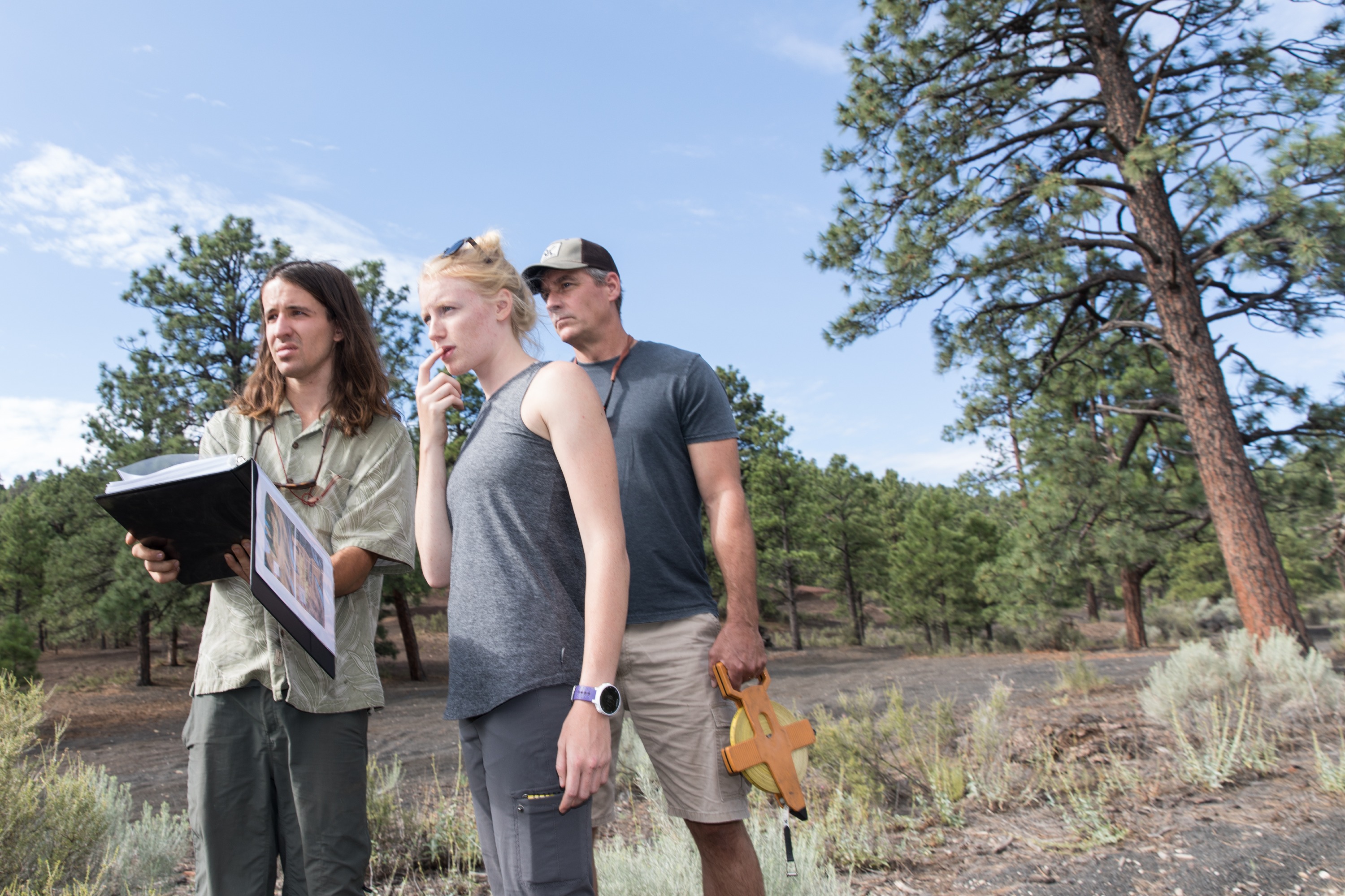 Three people standing in a wooded area with pine trees, examining a handheld device.