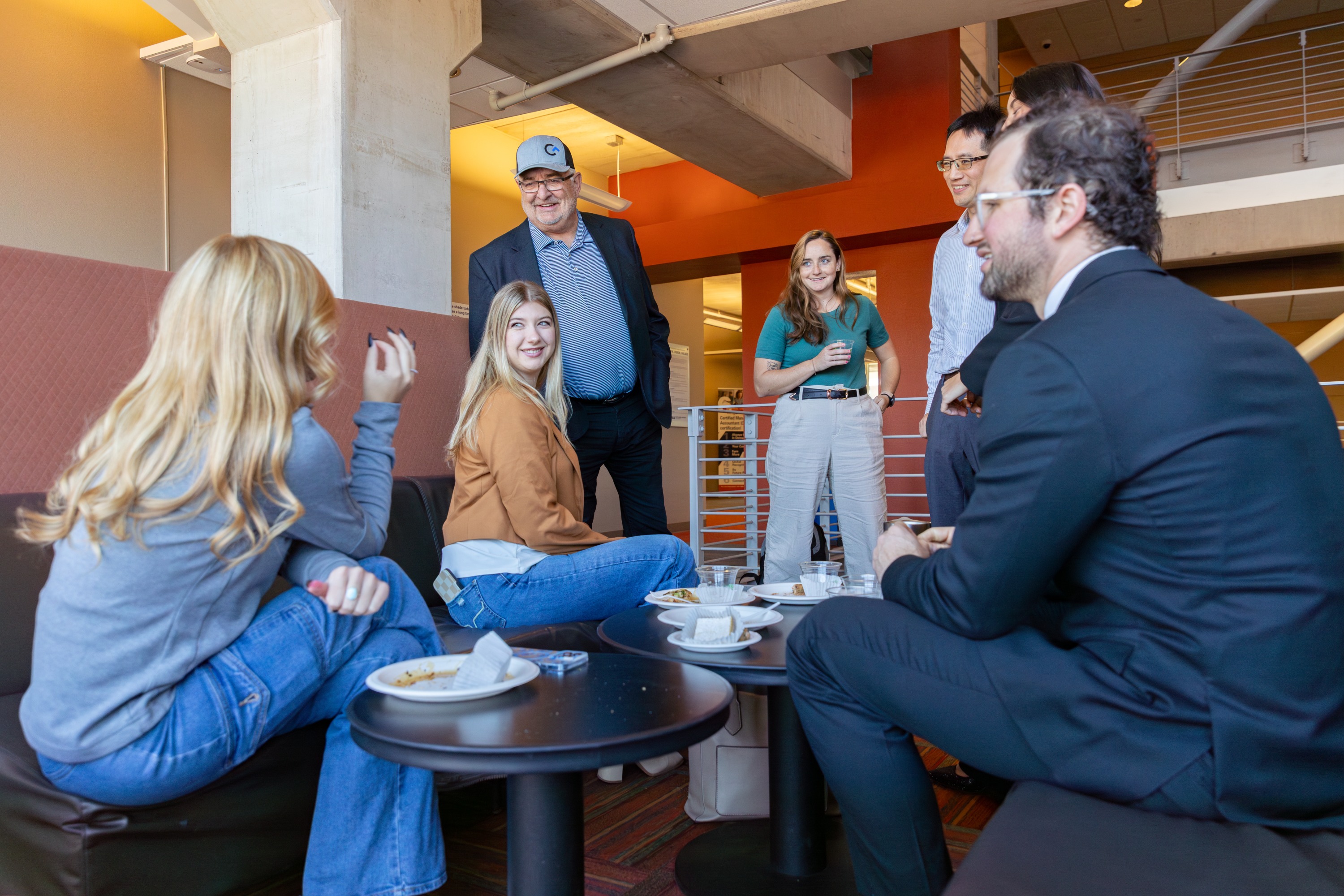 Alternative Text Six people gathered around a table in a casual dining setting. The people appear to be having a conversation while enjoying drinks and food. There are various items on the table including mugs and plates. The background features a warm and inviting decor with a combination of wood and warm colors.