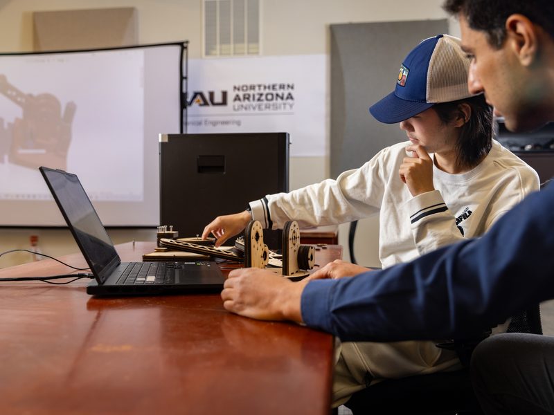 Two individuals sitting at a table, one using a laptop computer and the other observing. The laptop screen displays 'Northern Arizona University' text. The individuals are wearing casual clothing, including a baseball cap.