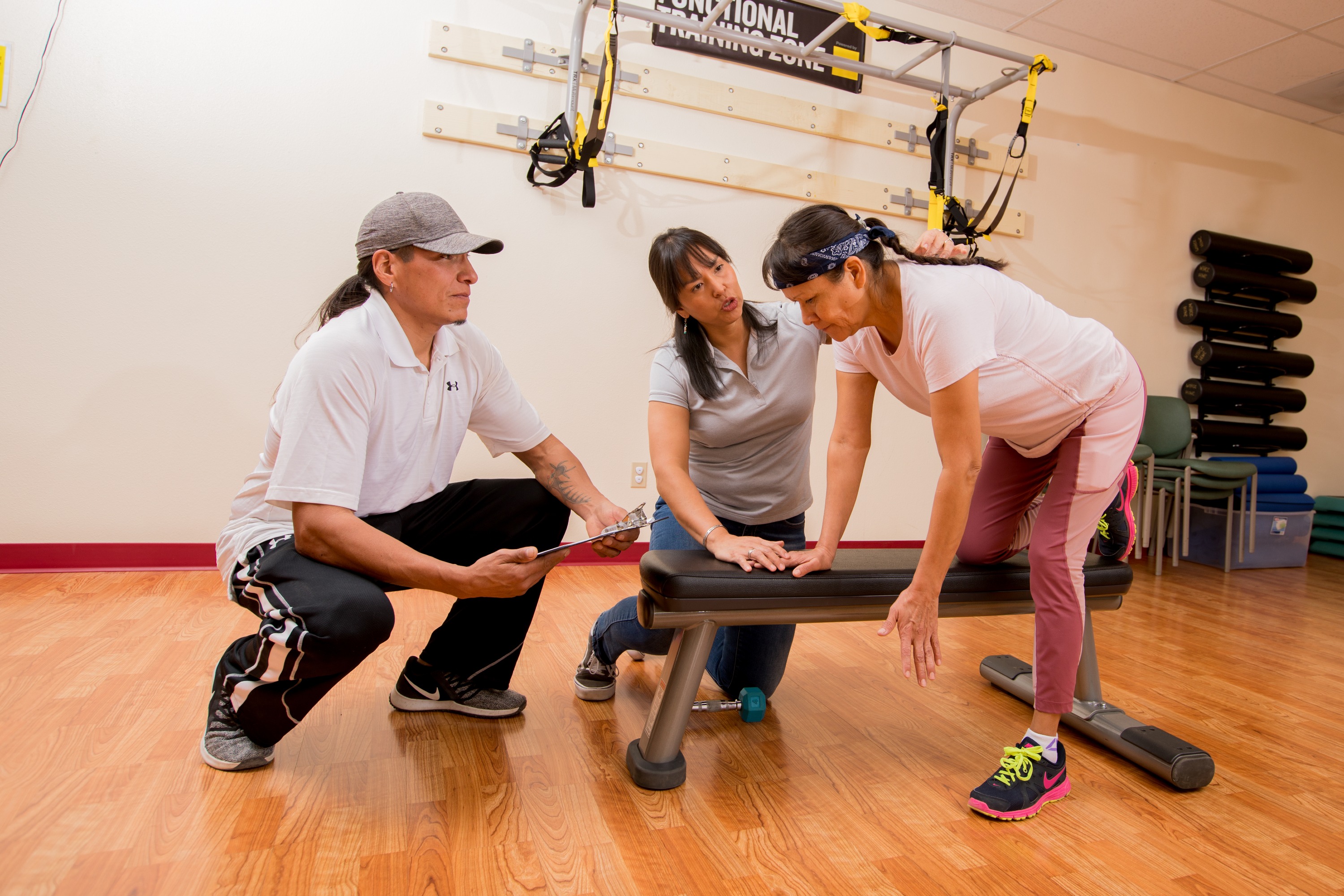 Three people, two females and one male, working together on exercise equipment in a fitness studio setting. There is various fitness equipment visible in the background, including resistance bands and a TRX suspension system.