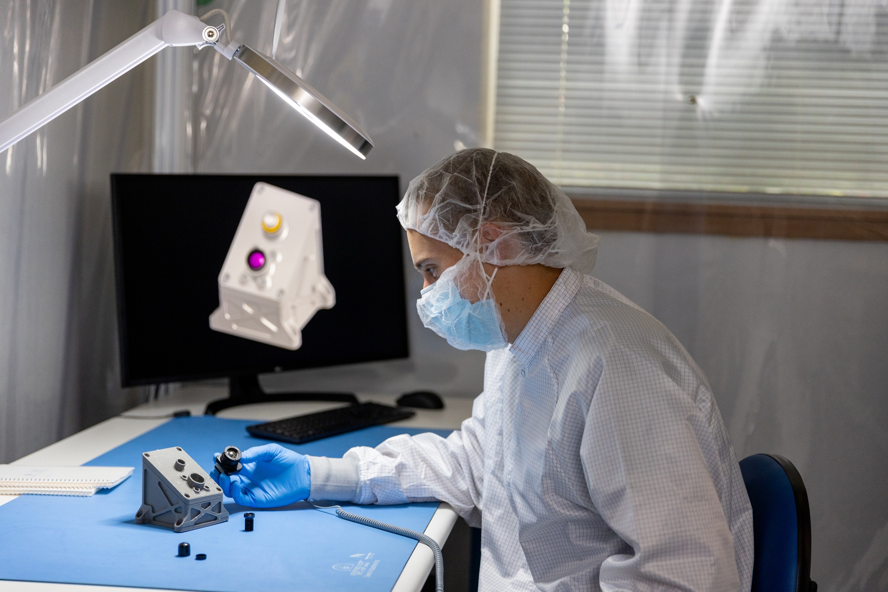 Alternative Text A person wearing protective medical gear, including a face mask, gloves, and a gown, working at a computer workstation in a laboratory or research setting. The person appears to be examining or working with a specimen displayed on the computer screen.