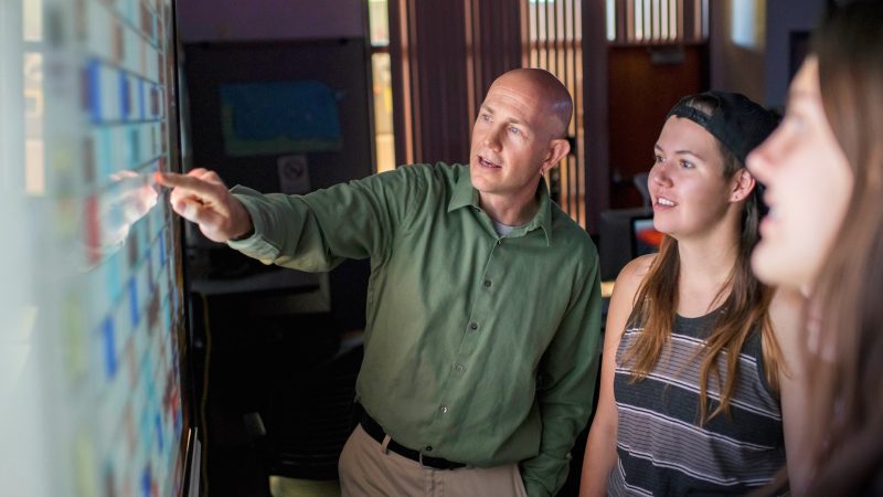Professor points to a screen while students watch.