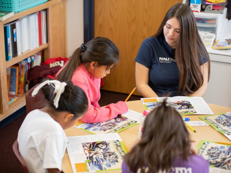 Student teacher sitting with children at a table, reading a book.