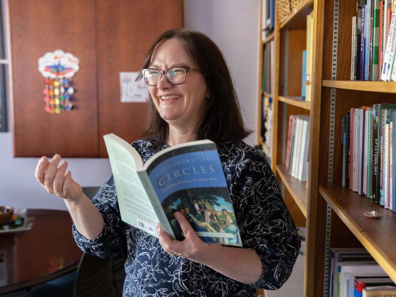 N A U faculty member reads book next to book shelf and smiles.