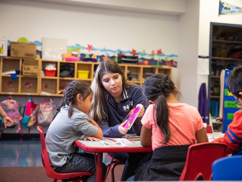 Student teacher helping elementary school kids with reading a book.