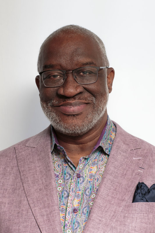 A professional portrait of a smiling man with short gray hair, a neatly trimmed gray beard, and rectangular glasses. He wears a light mauve blazer over a colorful paisley-patterned shirt, accented with a dark pocket square. He faces the camera against a plain white background, conveying warmth, confidence, and professionalism.