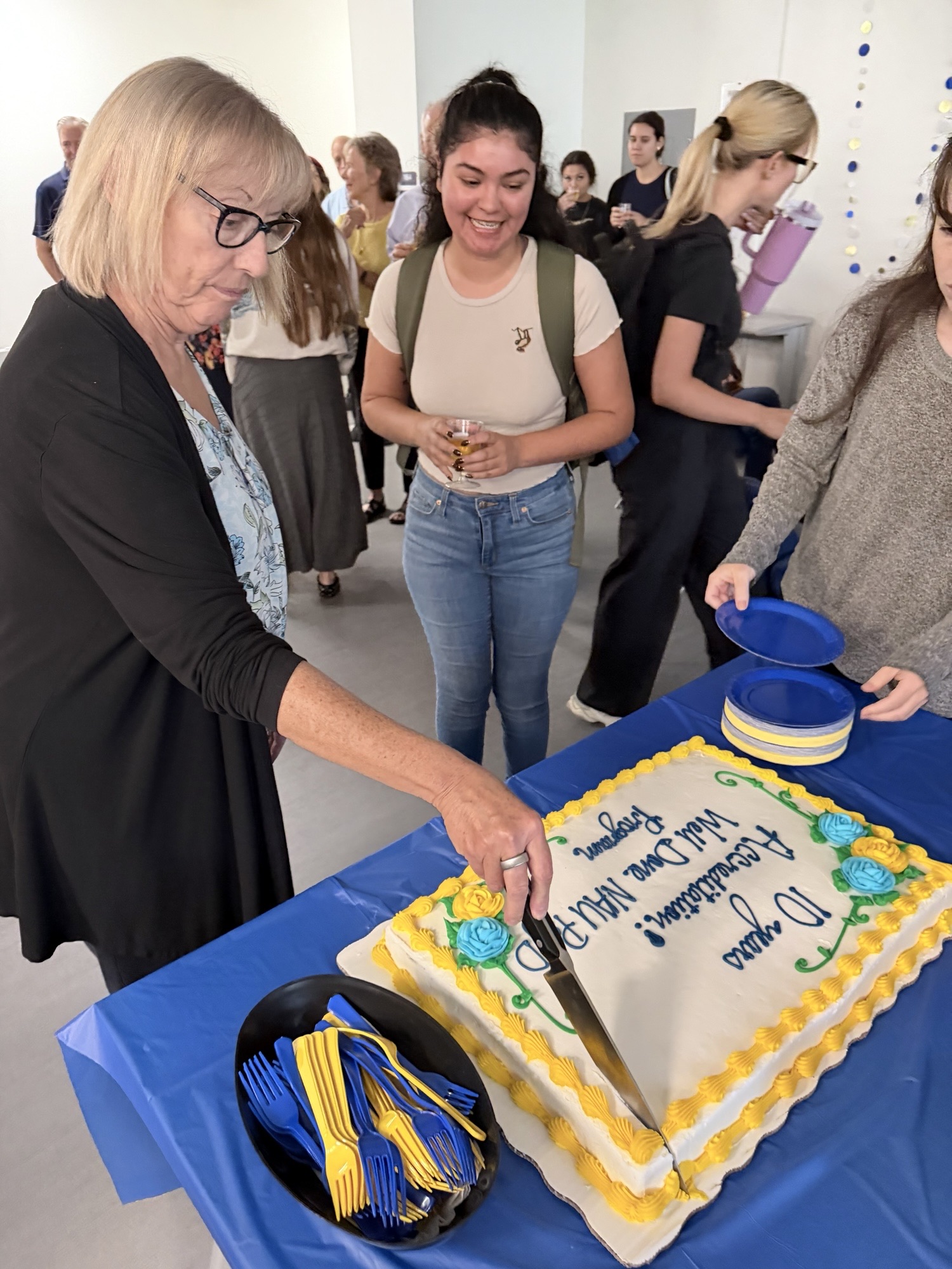A woman with short blonde hair and glasses cuts a large rectangular cake during a celebration marking 10 years of the Academic Psychology PsyD program. The cake is decorated with blue and yellow frosting, colorful flowers, and a congratulatory message. Students and colleagues gather around the table, smiling and watching. Blue and yellow plates and utensils complement the festive decorations, capturing a moment of community, celebration, and shared accomplishment.
