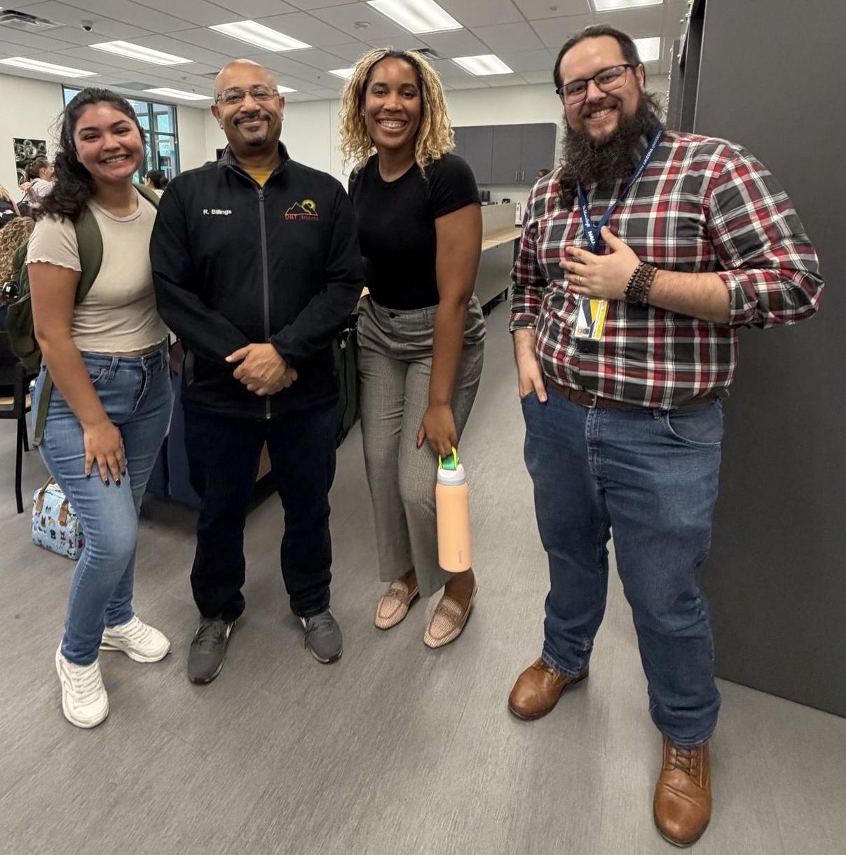 Four smiling individuals, including faculty and students, pose together in a bright university gathering space. Two women and two men stand side by side, dressed in casual and professional attire. One woman holds a peach-colored water bottle, while one man wears a university-branded jacket and another wears a plaid shirt with an identification badge. Other attendees socialize in the background near large windows, creating a welcoming atmosphere of connection and community.