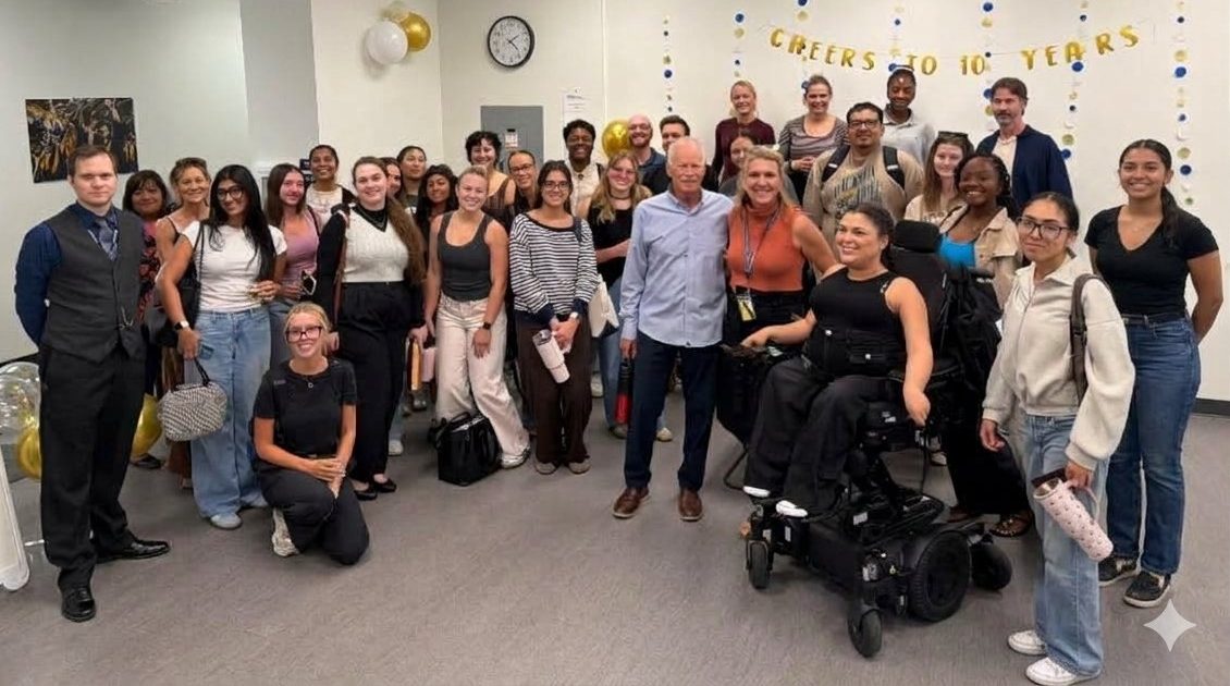 A large, diverse group of faculty and students poses together in a brightly lit indoor gathering space. Participants stand and kneel in several rows, smiling toward the camera, including a woman seated in a motorized wheelchair near the front. Blue, gold, and white decorations and a banner reading “Cheers to 10 Years” adorn the back wall, highlighting a celebratory milestone. The photograph captures a sense of community, inclusion, and shared accomplishment.