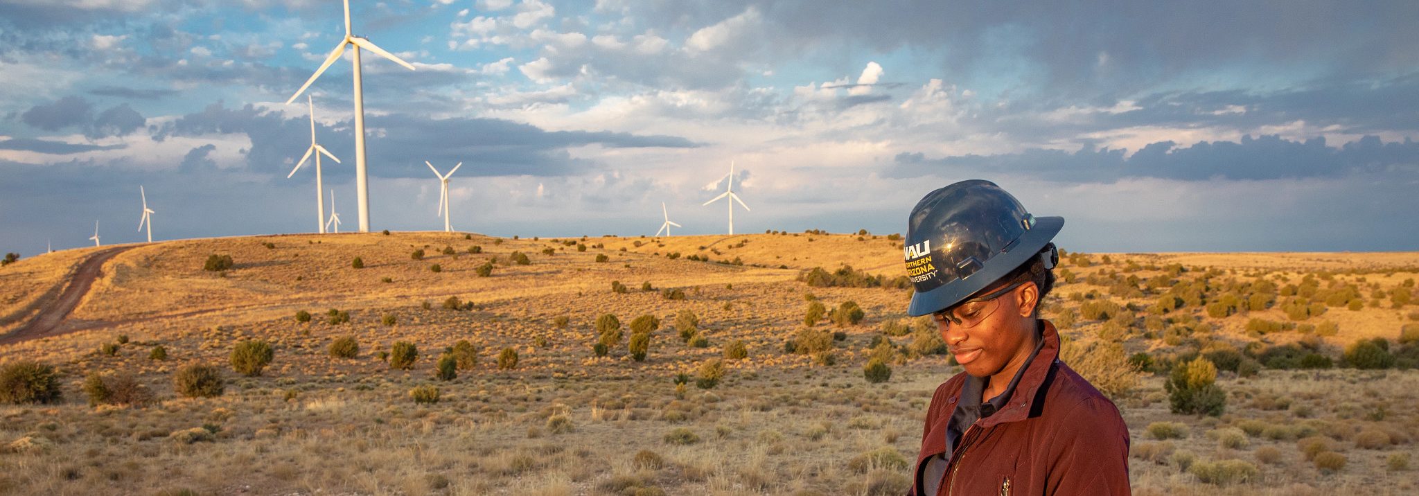A student wearing an N A U hardhat standing in a wind farm.