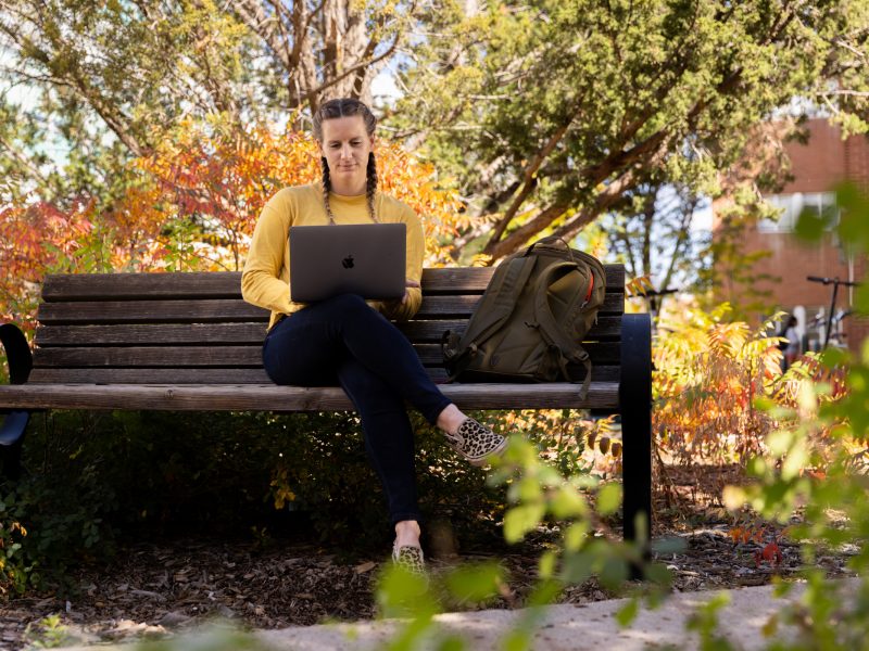 Female student working on a computer while sitting on a bench outside.