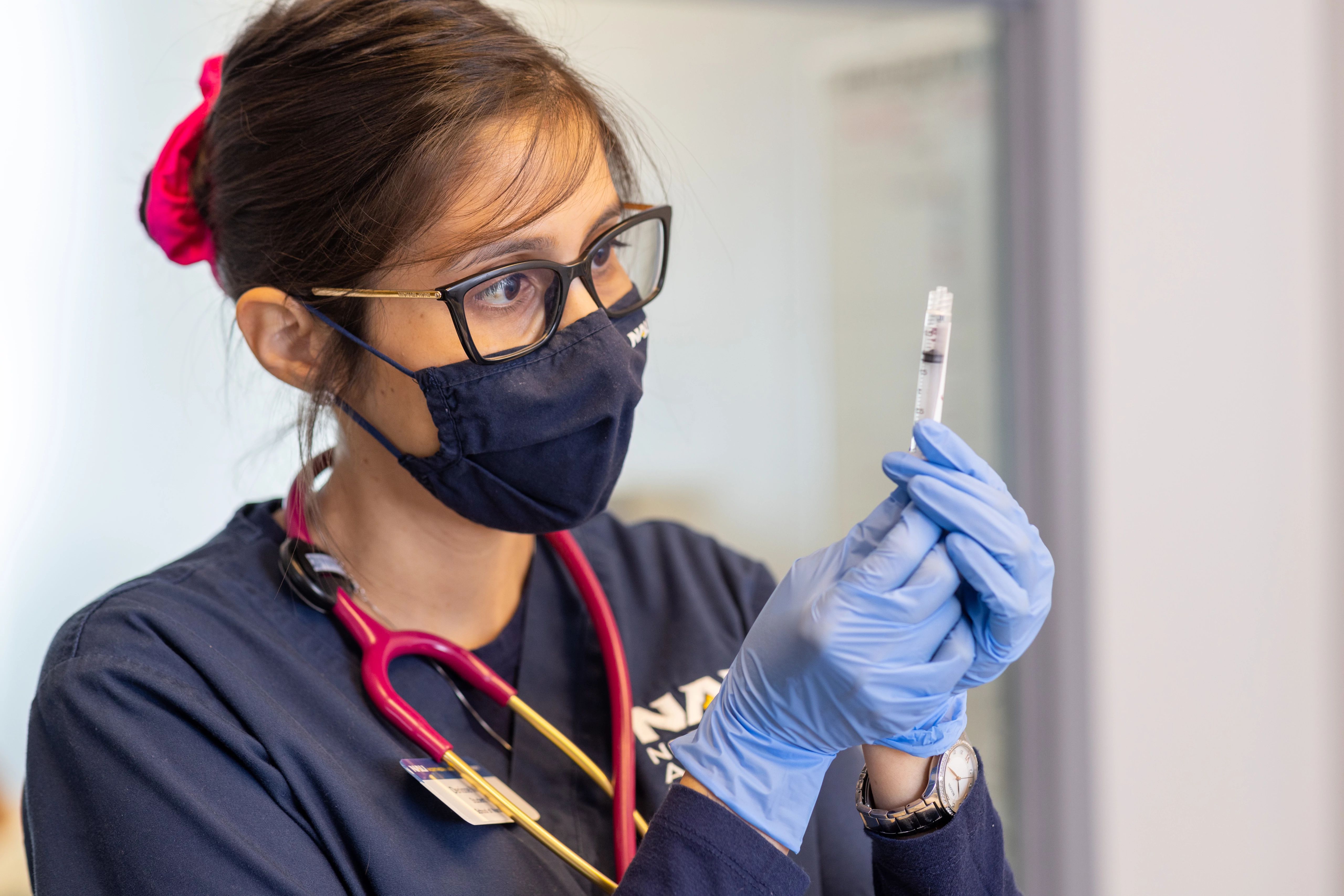 Nursing student wearing scrubs and medical mask, holding up a syringe.