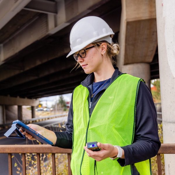 Civil Engineer major Jenna Hays, wearing a safety vest and hard hat, stands under bridge while looking at data on hand-held equipment.