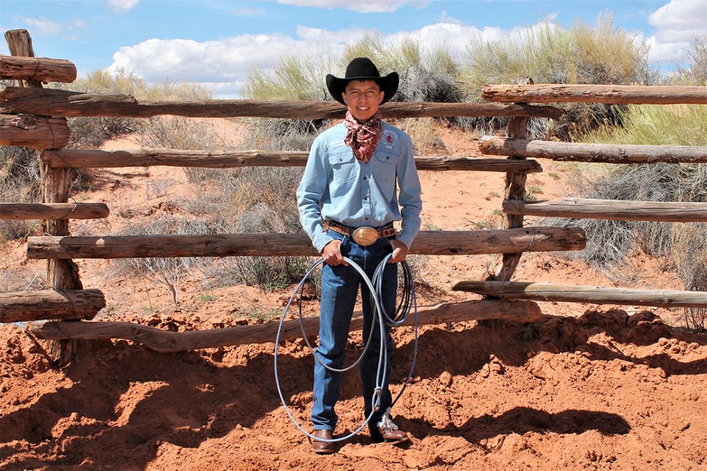Stade Riggs with a lasso and cowboy hat in front of a wood fence