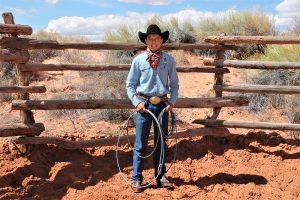 Stade Riggs with a lasso and cowboy hat in front of a wood fence