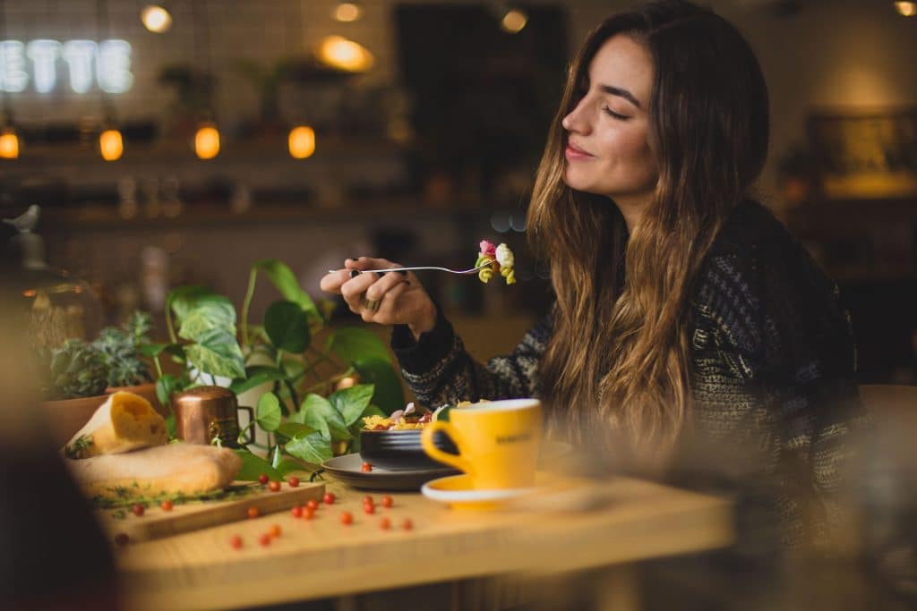 Woman about to take bite of pasta with her eyes closed showing eating mindfully. The woman is eating a table with a coffee cup on it, a vine of leaves going across it and cranberries scattered on it.