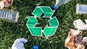 Looking down with grass as the background the green recycilng sign with three curving arrows following each other in a triangle shape is imposed in the middle of the image. Pieces of recyclable materials like water bottles and paper lay around the sign