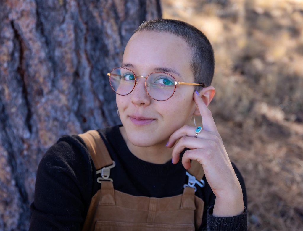 Portrait of Sydney Rittershaus wearing overalls and a black shirt with her hand to her face