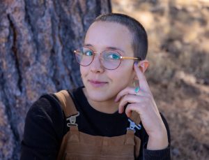 Portrait of Sydney Rittershaus wearing overalls and a black shirt with her hand to her face