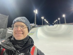 Ian McLeod wearing a Medicine in Motion beanie and posing in front of a snowboard halfpipe course in Aspen, Colorado