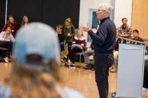 Man with gray hair speaking to students sitting around the edges of a classroom
